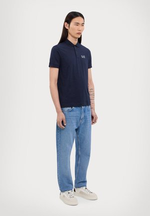 Navy blue polo shirt with short sleeves, featuring a collar and an embroidered logo. Paired with light blue jeans and beige sneakers.