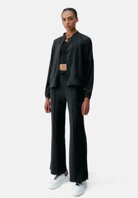 Completo atletico nero composto da una giacca bomber con zip frontale, un top corto e pantaloni a gamba larga, tutti realizzati in un tessuto liscio e opaco. Sneakers bianche.