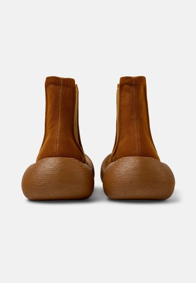 Bottines en daim marron avec une semelle en caoutchouc ronde, présentant une finition texturée et des panneaux latéraux en élastique pour un enfilage facile.