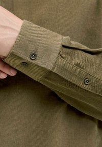 Primo piano di una mano che regola il polsino di una camicia in velluto a coste marrone con bottoni visibili e tessuto testurizzato.