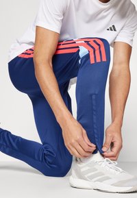 Persona ceļos, aizverot zilas sporta bikses ar sarkaniem svītrām pie potītes, valkājot baltas sporta kurpes un balto kreklu.