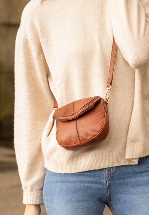 Brązowa skórzana torebka crossbody o zaokrąglonym kształcie, ze złotym zamkiem błyskawicznym i krótkim paskiem, noszona na jasnoszarym swetrze i niebieskich dżinsach.