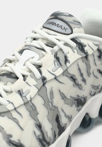 Zapatilla blanca y gris con patrón, con parte superior de malla, que presenta un diseño texturizado y lengüeta acolchada, complementada por suelas claras y redondeadas.