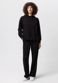 Sudadera negra con cordones ajustables, corte relajado, bolsillo frontal; combinada con pantalones de pierna ancha negros y zapatillas blancas.