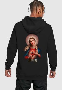 Felpa nera con cappuccio caratterizzata da una grafica colorata di una donna che tiene un cuore, con un'aureola e la parola "PRAY" in oro sotto l'immagine.