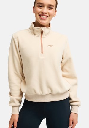 Personne souriante portant un pull polaire beige avec une fermeture éclair quart et un pantalon noir, debout devant un fond blanc.