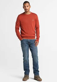 Maglione rosso a maglia con polsini e fondo a costine, con un piccolo logo. Abbinato a jeans in denim blu e scarpe marroni.