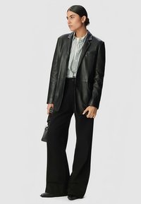 Zwarte leren blazer over een witte en gestreepte blouse, gecombineerd met zwarte broek met wijde pijpen, en een kleine, gestructureerde zwarte handtas in de hand.