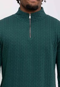 Pull vert à col zippé avec un motif tricoté texturé, en tissu léger et à coupe ajustée avec des éléments de fermeture éclair visibles.
