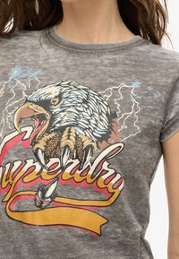 Šedé grafické tričko s designem orla, který má podrobné peří a drápy, spolu s textem "Superdry" v červených a žlutých akcentačních barvách.