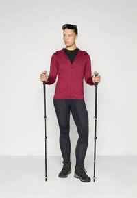 Roter Kapuzenpullover mit Reißverschluss, schwarzes Shirt, schwarze Leggings mit strukturierten Akzenten. Hält Wanderstöcke und trägt robuste schwarze Wanderschuhe.