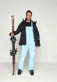 Skiausrüstung mit schwarzer Jacke, hellblauen Skihosen und schwarzen Skischuhen, die Skier mit Bindungen hält. Einfaches Design mit funktionalen Elementen.