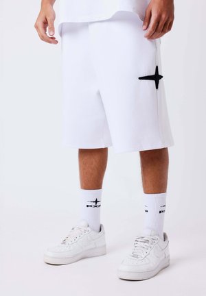 Persoon draagt witte lange shorts met zwart kruislogo, bijpassende witte sokken met logo en witte Nike-sneakers tegen een effen achtergrond.