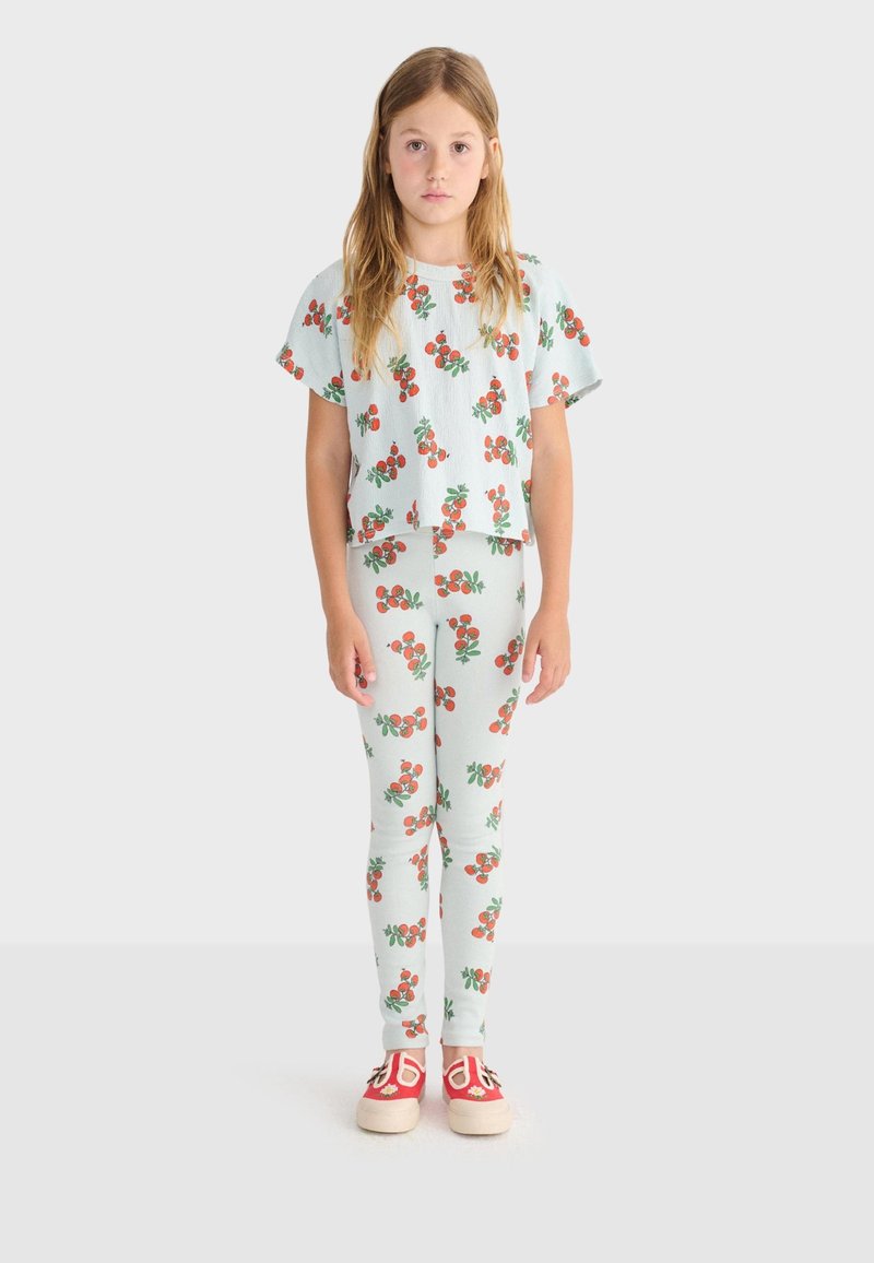 Jeune fille aux longs cheveux portant une tenue blanche assortie avec des motifs de cerises rouges et des chaussures rouges à enfiler, debout devant un fond uni.