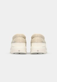 Zapatillas beige acolchadas con una suela texturizada blanca. Presentan un acabado suave y un detalle sutil de logo en el lado exterior.