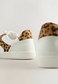 White sneakers with a tan leopard-print accent at the heel, textured upper, and contrasting smooth rubber sole. Rounded toe design.