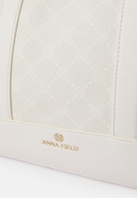 Borsa bianca con un delicato motivo a logo, realizzata in morbida ecopelle e dotata di hardware dorato con il dettagli embossato "ANNA FIELD".