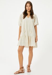 Cremfarbnes Kleid mit kurzen Puffärmeln, einem V-Ausschnitt und einem gewellten Saum. Verziert mit einem Spitzenmuster und Quasten am Kordelzug. Getragen mit Sandalen.