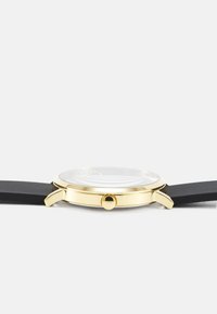 Runder Uhr mit einem Gehäuse aus goldfarbigem Edelstahl, einem schwarzen Silikonarmband und einem glatten Glaszifferblatt mit einem filigranen Design darunter.