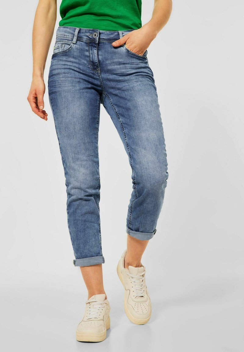 Světle modré úzké džíny s ohrnutými nohavicemi, pěti kapsami a jemným vyblednutím, v kombinaci se krémovými teniskami na jednobarevném pozadí.