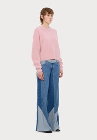 Άτομο που φοράει ανοιχτό ροζ χνουδωτό πουλόβερ και ψηλόμεσα παντελόνια με φαρδιά μπατζάκια και τριγωνικά ανοιχτόχρωμα τζιν μπαλώματα στο τελείωμα.