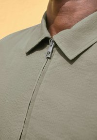 Olivovo zelená košeľa s textúrovanou látkou, ktorá má plochý golier a predné zapínanie na zips. Golier je štrukturovaný a zips je kovový.