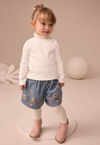White turtleneck top, blue corduroy shorts with floral embroidery, white textured tights, and pink ankle boots. Child standing on a circular base.