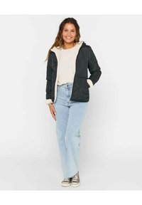 Veste noire matelassée avec doublure en polaire crème, manches longues et poches, portée sur un pull léger et un jean bleu. Coupe décontractée avec un design simple.
