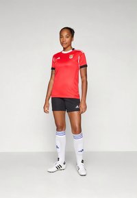 Kvinne som står iført en rød og svart fotballuniform med hvite knelange sokker og fotballsko på en lys grå bakgrunn.