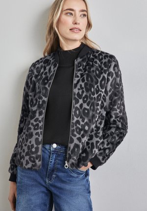 Giacca bomber con stampa leopardata, in pelle sintetica nera, chiusura a zip, polsini e colletto a costine. Indossata sopra un top in maglia nera e jeans blu.