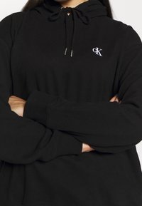 Czarna bluza z kapturem, wykonana z miękkiego materiału, z kieszenią typu kangurka, ściągaczem na kapturze, prążkowanymi mankietami oraz małym białym logo CK na klatce piersiowej.