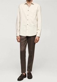 Uomo che indossa una camicia a maniche lunghe color crema con bottoni, pantaloni marroni su misura e mocassini marroni scuri, in piedi contro uno sfondo neutro.