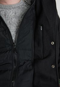 Schwarze Jacke mit einem Reißverschluss vorne, ausgestattet mit einem gesteppten schwarzen Innenfutter und einem grauen Strickpullover darunter, hervorgehoben durch ein strukturiertes Muster.