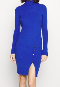 Ribbad stickad klänning i klargul. Den har en hög halsringning, långa ärmar, en sidofåra och guldknappdekorationer längs vänstra sömmen.