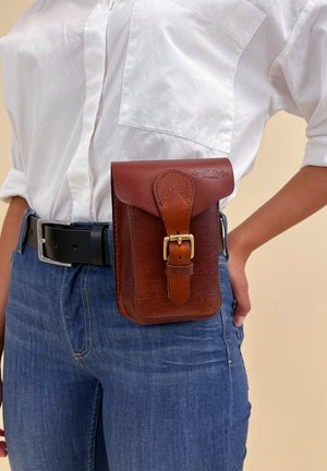 Person trägt blaue Jeans, weißes Hemd und schwarzen Gürtel mit einer braunen Leder-Gürteltasche, die am Taillenbereich durch eine goldene Schnalle gesichert ist.