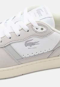 Zapatillas Lacoste en una combinación de cuero gris claro con acentos blancos, con cordones blancos y un logo de cocodrilo cerca del talón.