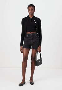 Svart ribbet cardigan med rund krage og knappelukking over en svart crop top, kombinert med svarte høytlivde denimshorts og flate sko.
