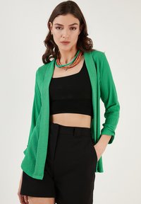 Zelený, textúrovaný kardigán s otvoreným predným dielom a dlhými rukávmi, nosený cez čierny cropped top a čierne šortky. Doplnky zahŕňajú vrstvené náhrdelníky.