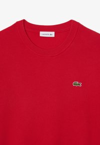 Pull rouge à col rond avec col côtelé et petit logo crocodile vert Lacoste brodé sur le côté gauche de la poitrine.