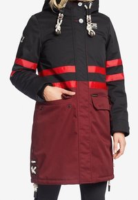 Lange parka jas met een zwart bovenwerk, rode gestreepte accenten en een bordeauxrode ondersectie. Inclusief een capuchon, trekkoord en voorzakken.