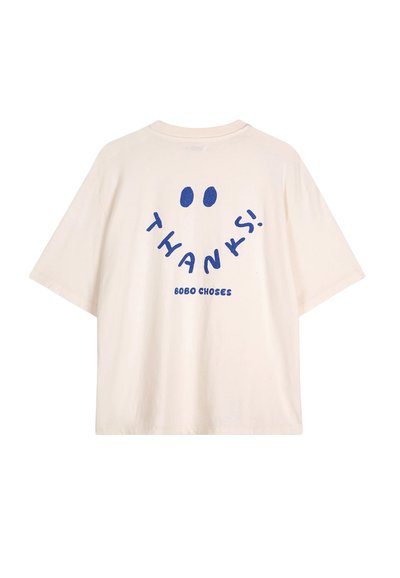 Camiseta de algodón color crema con un corte relajado. Presenta el texto azul "¡GRACIAS!" y un diseño de carita sonriente en la parte posterior, con el nombre de la marca impreso.