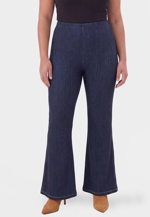Person, die dunkelblaue ausgestellte Jeans und braune spitz zulaufende Stiefel mit Absatz trägt, steht mit entspannt an den Seiten hängenden Armen.