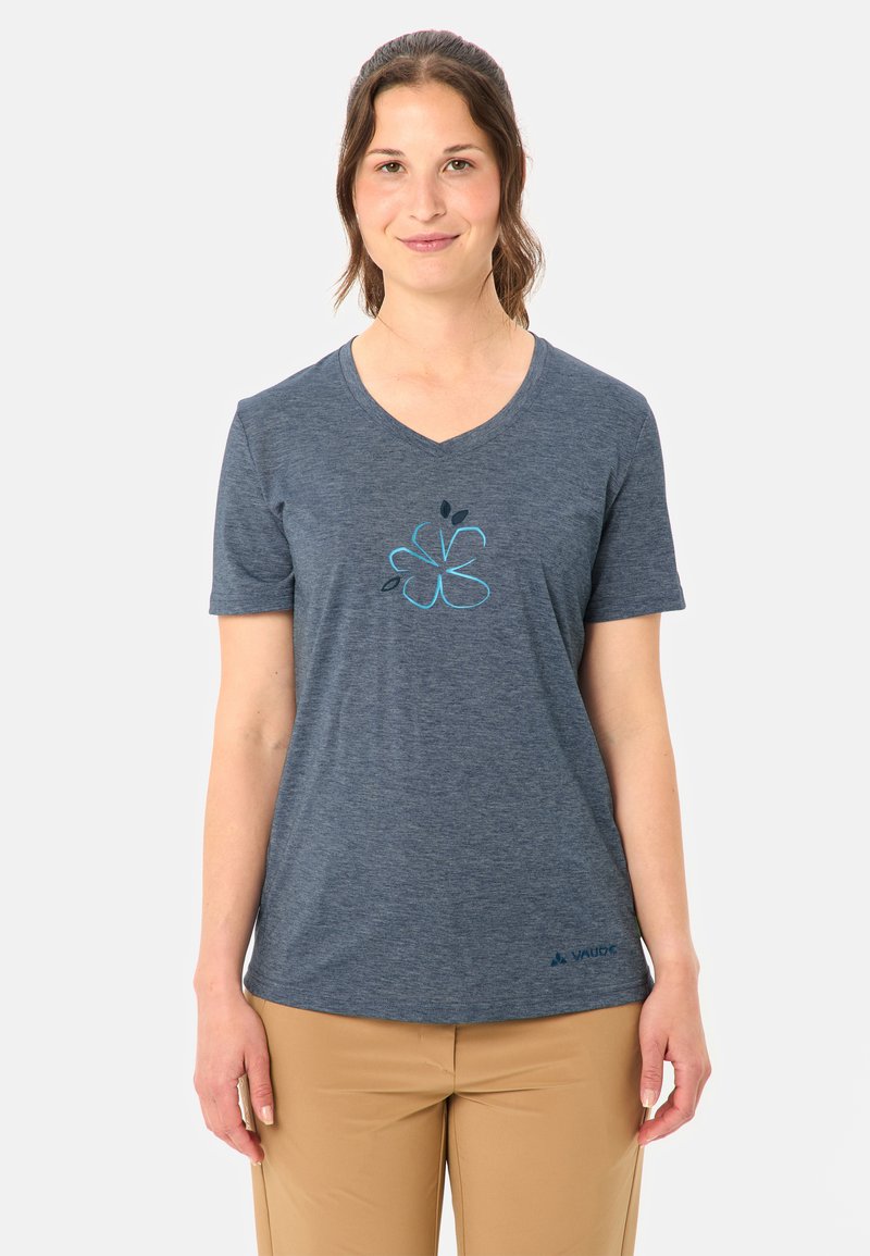 Femme aux cheveux bruns portant un T-shirt bleu à col en V avec un motif floral et un pantalon beige, debout devant un fond blanc uni.