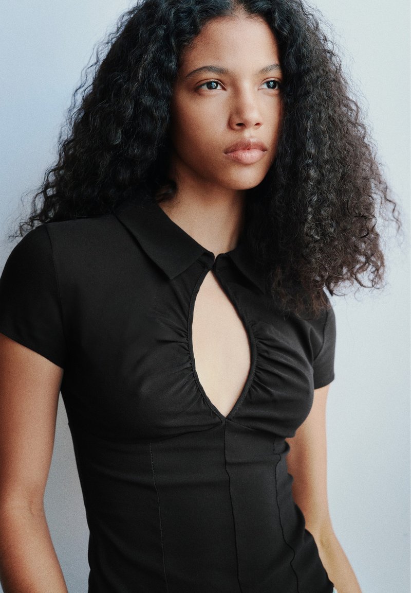Jeune femme aux cheveux bouclés portant une robe noire cintrée avec un décolleté en trou de serrure, regardant légèrement vers la droite sur un fond clair uni.