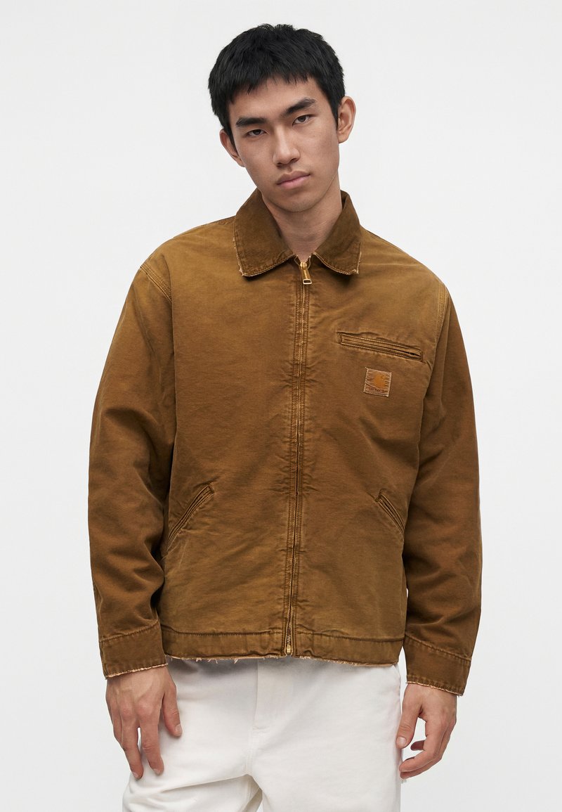 Junger Mann, der eine braune Jacke mit Reißverschluss und Kragen und weiße Hosen trägt, steht vor einem schlichten hellen Hintergrund.
