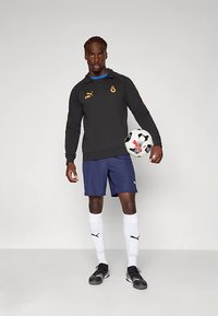 Fekete sport pulóver narancssárga díszítéssel, sötétkék rövidnadrág, fehér zokni és fekete stoplis cipő. Egy fekete-fehér focilabdát tart.