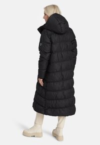 Naine, kes kannab pikka musta sulejope, millel on kapuuts, kreemivärvi pükse ja beeži nööbiga jalanõude komplekti, seisab ja vaatab kõrvale.