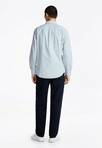 Blauw-wit gestreepte langmouw shirt met een button-down kraag, gecombineerd met donkere broek en witte schoenen. Gezicht vanuit de achterkant.