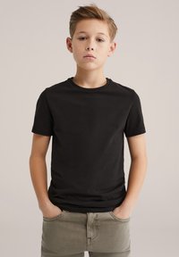 Fekete pamut póló rövid ujjakkal és kerek nyakkivágással, világoszöld nadrággal párosítva. Egyszerű kialakítás sima textúrával, látható minták nélkül.