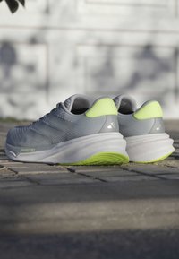 Baskets de sport grises avec une tige texturée, des accents verts au niveau du talon et une semelle blanche. Présente un design épuré et une semelle à rembourrage prononcé.